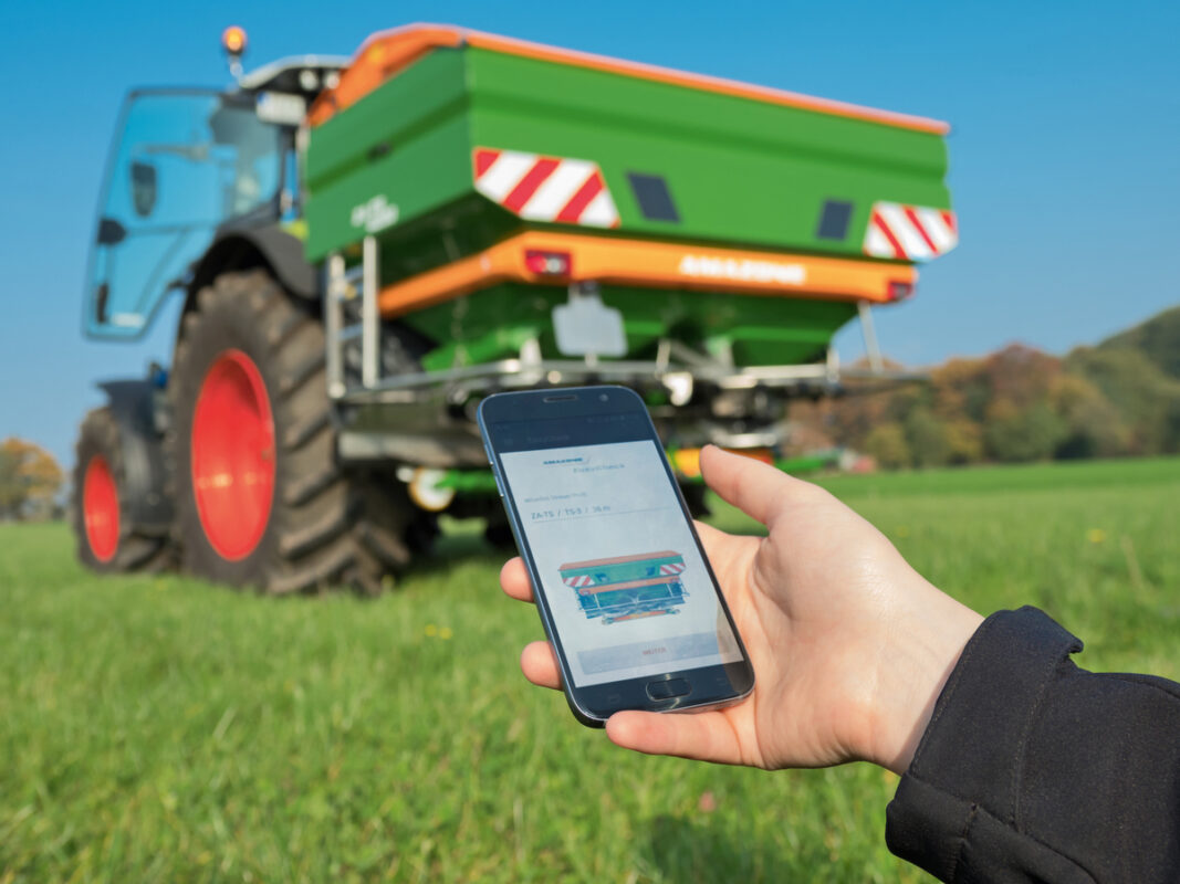 Un tracteur avec un épandeur d'engrais et une main qui tient un smartphone avec l'application mobile pour les réglages de l'épandeur