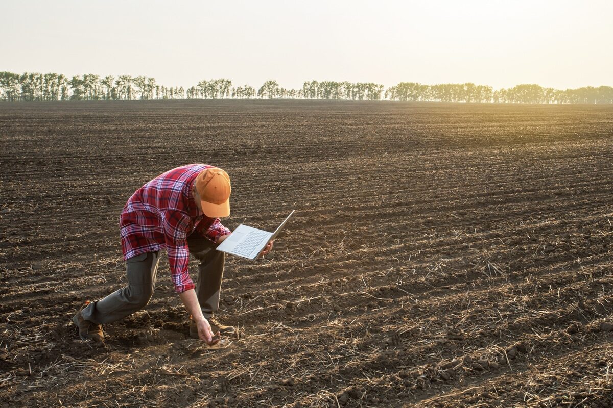 un homme baissé dans un champ labouré avec un ordinateur à la main - Illustration Anticiper les démarches environnementales et bâtiment