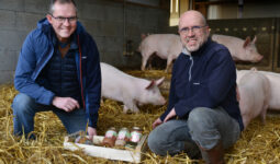 2 hommes présentant des bocaux de plats cuisinés avec des porcs élevés sur paille derrière eux